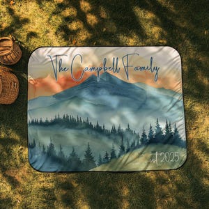 May include: A rectangular picnic blanket with a watercolour mountain landscape in blue, green, and orange hues. The text "The Campbell Family" is above the mountains, with "est 2025" below. The blanket is designed for outdoor use.