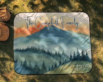 May include: A personalized picnic blanket featuring a watercolor mountain scene in shades of blue, green, and orange. The text "The Campbell Family" is displayed above the mountain range, with "est 2025" below. The blanket is rectangular.