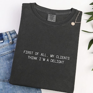 May include: Dark grey Comfort Colors t-shirt with the text "FIRST OF ALL, MY CLIENTS THINK I'M A DELIGHT" in white. A gold necklace with a pendant is pinned to the shirt. Blue jeans are visible in the background.
