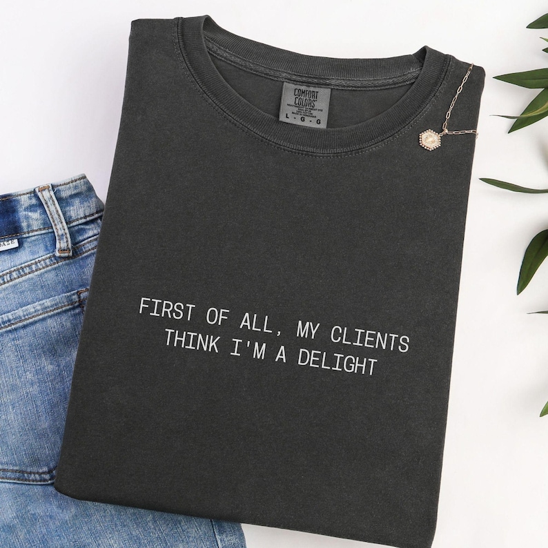 Puede incluir: Camiseta Comfort Colors gris oscuro con el texto "FIRST OF ALL, MY CLIENTS THINK I'M A DELIGHT" en blanco. Un collar dorado con un colgante est&aacute; prendido a la camiseta. Se ven unos vaqueros azules en el fondo.