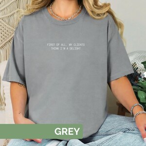 Puede incluir: Una camiseta gris con el texto "FIRST OF ALL, MY CLIENTS THINK I'M A DELIGHT" en blanco. La palabra "GREY" se muestra en un rect&aacute;ngulo verde en la parte inferior de la camiseta.