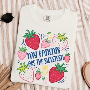 Może przedstawiać: Biała koszulka z kolorowym wzorem truskawek i napisem "MY PATIENTS ARE THE SWEETEST!". Truskawki są czerwone, różowe i żółte z zielonymi liśćmi. Koszulka jest złożona na paskowanej powierzchni.