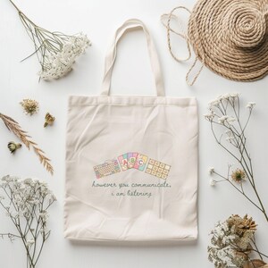 May include: Cream-colored tote bag with a colorful keyboard and ABC design. The bag has a quote that reads "however you communicate, I am listening." The bag is surrounded by dried flowers and a straw hat.
