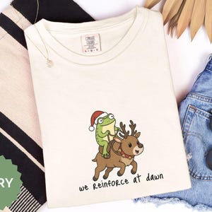 May include: An ivory t-shirt featuring a graphic of a green frog wearing a red and white Santa hat, riding a brown reindeer with a red collar and gold bells. Below the graphic, the text reads, we reinforce at dawn in black lettering.