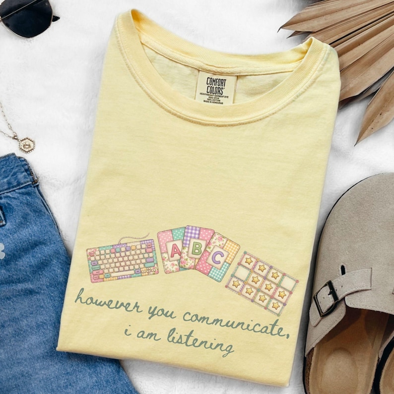May include: A light yellow t-shirt with a graphic of a keyboard, ABC cards, and a star-shaped grid. The text "however you communicate, i am listening" is printed below the graphic. The shirt is made of a soft material.