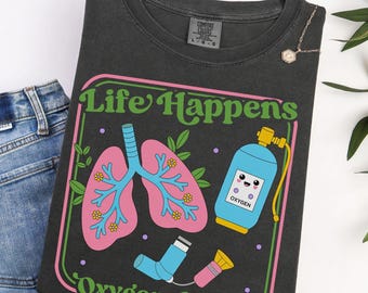 Puede incluir: Una camiseta gris carbón con un gráfico que incluye el texto Life Happens Oxygen-Helps. El diseño muestra pulmones rosas con bronquios azules y flores amarillas, un tanque de oxígeno azul con una cara y un inhalador azul y rosa.