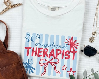 May include: White t-shirt with the words "occupational THERAPIST" in red, blue, and white. The design includes stars, stripes, and a bow. The shirt is made of a soft material and is a Comfort Colors brand.