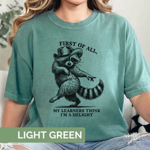 May include: Light green t-shirt featuring a raccoon in a cowboy hat with the text "FIRST OF ALL, MY LEARNERS THINK I'M A DELIGHT". The shirt is light green in colour.