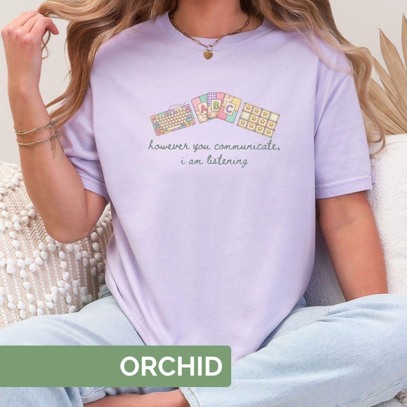 May include: Orchid-colored t-shirt with a graphic of keyboards and ABC blocks. The text "however you communicate, I am listening" is printed below the graphic. The word "ORCHID" is printed in a green rectangle at the bottom.