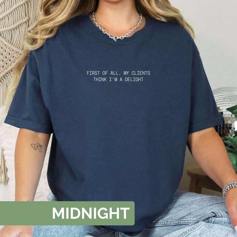 Puede incluir: Una camiseta azul marino con el texto "FIRST OF ALL, MY CLIENTS THINK I'M A DELIGHT" en blanco. La palabra "MIDNIGHT" se muestra en un rect&aacute;ngulo verde en la parte inferior de la camiseta. La camiseta tiene mangas cortas y un corte holgado.