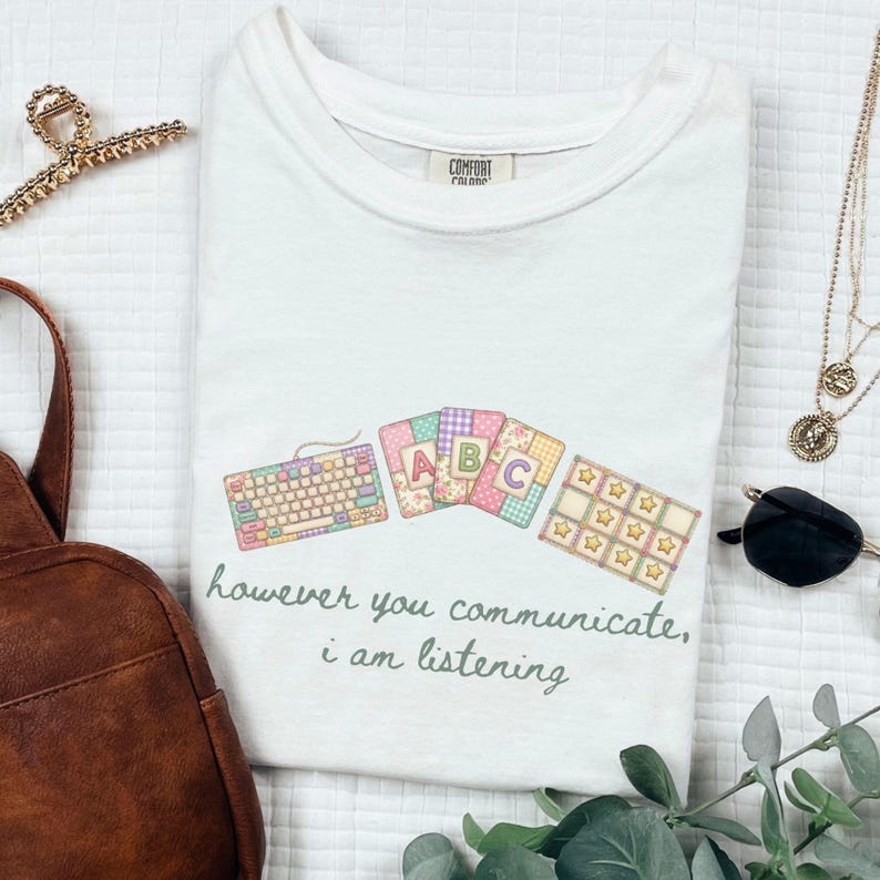 May include: White t-shirt with a colorful graphic featuring a keyboard, ABC blocks, and star-shaped elements. The text "however you communicate, i am listening" is printed below the graphic. The shirt is a Comfort Colors brand.