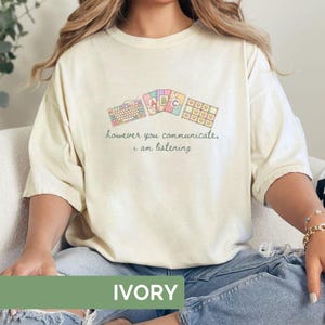 May include: Ivory-colored t-shirt with a whimsical design featuring keyboard and ABC graphics. The shirt has the text "however you communicate, i am listening" in a soft green color. The shirt is made of a soft material.
