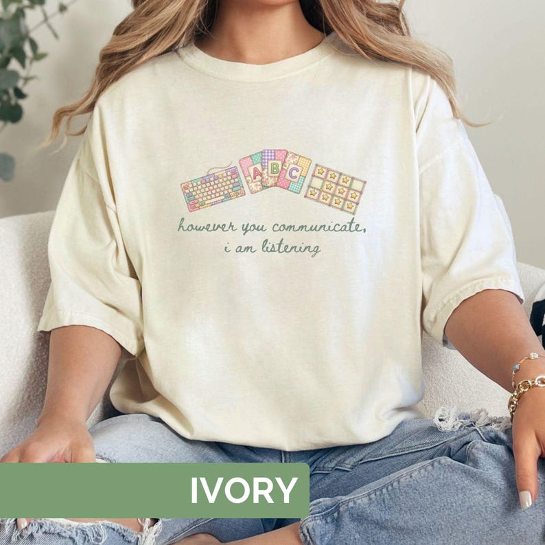 May include: Ivory-colored t-shirt with a whimsical design featuring keyboard and ABC graphics. The shirt has the text "however you communicate, i am listening" in a soft green color. The shirt is made of a soft material.