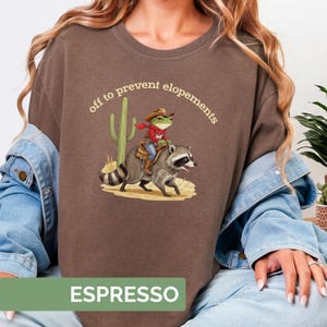 Könnte beinhalten: Espressofarbenes Sweatshirt mit einer Grafik einer Frosches mit Cowboyhut, der auf einem Waschbären reitet, mit einem Kaktus im Hintergrund. Der Text "off to prevent elopements" ist über der Grafik angeordnet. Das Wort "ESPRESSO" steht unten.