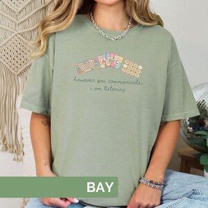 May include: Sage green t-shirt with a colorful graphic of keyboard keys and ABC blocks. The text "however you communicate, i am listening" is printed below the graphic. The word "BAY" is printed in a green rectangle at the bottom of the image.