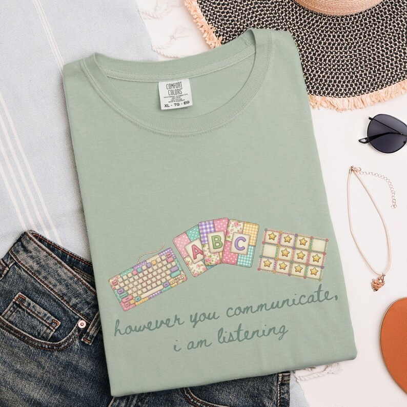 May include: Sage green t-shirt with a graphic featuring a keyboard, ABC cards, and star-shaped blocks. The text "however you communicate, i am listening" is printed below the graphic. The shirt is a Comfort Colors XL-TG-EG.