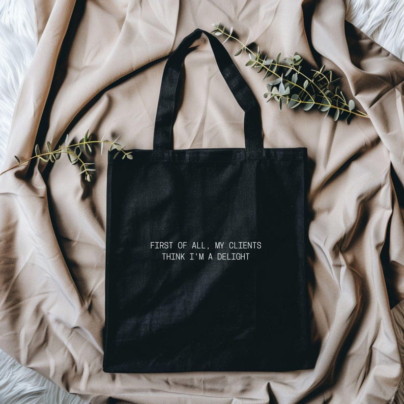 Puede incluir: Bolso tote negro con el texto "FIRST OF ALL, MY CLIENTS THINK I'M A DELIGHT" en blanco. El bolso tiene dos asas negras y se muestra sobre una tela beige con vegetaci&oacute;n.