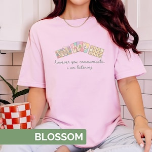 May include: A light pink t-shirt with the word "BLOSSOM" in a green rectangle at the bottom. The shirt features a keyboard and ABC blocks graphic with the text "however you communicate, i am listening."