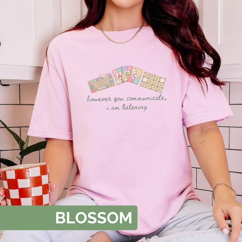 May include: A light pink t-shirt with the word "BLOSSOM" in a green rectangle at the bottom. The shirt features a keyboard and ABC blocks graphic with the text "however you communicate, i am listening."