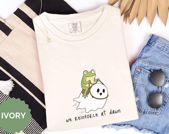 Puede incluir: Una camiseta de color marfil con un gráfico de una rana verde montada sobre un fantasma blanco. La rana sujeta unas riendas marrones atadas al fantasma. Debajo de la ilustración, el texto "we Reinforce at dawn" aparece en letras negras.