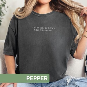 Puede incluir: Una camiseta gris oscuro con el texto blanco "FIRST OF ALL, MY CLIENTS THINK I'M A DELIGHT". La palabra "PEPPER" se muestra en un rect&aacute;ngulo verde en la parte inferior de la camiseta.
