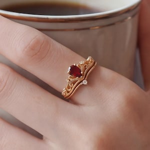 May include: A gold ring featuring a pear-shaped deep red gemstone at the center, surrounded by intricate filigree metalwork and small clear accent stones. The band has a curved, crown-like design that sits prominently on the finger.