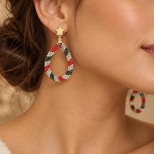 Pode incluir: Um par de brincos pendentes com um pino em forma de estrela dourada ligado a um aro em forma de gota. O aro é coberto por pequenas contas alternadas em vermelho, verde e branco, criando um padrão de riscas em toda a estrutura circular.