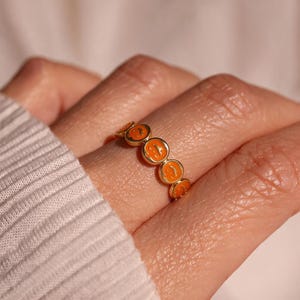 Puede incluir: Un anillo de tono dorado que presenta una serie de pequeños motivos circulares de calabaza en esmalte naranja dispuestos en una banda continua. Cada diseño de calabaza incluye detalles tallados que representan ojos y boca.