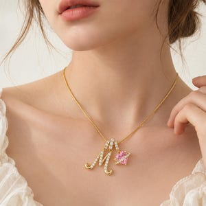 Puede incluir: Un collar de cadena dorada con un colgante en forma de letra M cursiva incrustado con piedras blancas transparentes y un pequeño dije en forma de corazón rosa rodeado por un anillo dorado, suspendidos de una fina cadena de metal dorado.