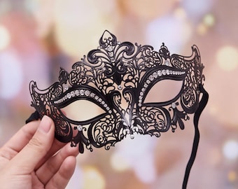 May include: A black masquerade mask featuring intricate filigree metalwork and clear rhinestones embedded along the eye openings and the central crest. The mask has a pointed top design and includes a thin black elastic strap for secure attachment.