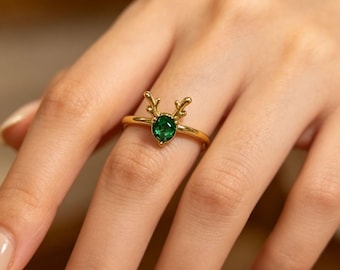 May include: A gold ring featuring a central oval-shaped green gemstone. The setting is designed with two small, branching antler-like prongs extending upward from the base of the stone, securing it within the polished gold band.