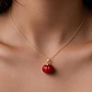 May include: A gold chain necklace featuring a small, round, red pendant with vertical ridges. The pendant is suspended from a simple gold bail attached to a delicate gold link chain that rests against the collarbone area of the neck.