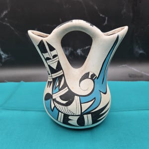 May include: A handcrafted, off-white ceramic vase with a handle. The vase features a design with black and turquoise geometric shapes. The vase is set against a turquoise background.