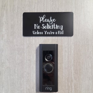 May include: A black doorbell camera with the word "ring" printed on the bottom. A sign above the camera reads "Please No Soliciting Unless You're a Kid."