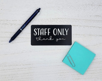 以下が含まれることがあります： 白い文字で「STAFF ONLY thank you」と書かれた黒い看板。青いペンと銀色のクリップが付いたターコイズブルーの付箋が白い木製の表面にある。
