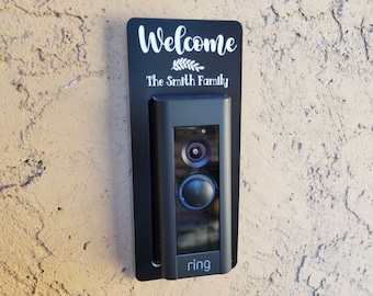 以下が含まれることがあります： WelcomeとThe Smith Familyという白い文字と小さな葉のグラフィックが描かれた黒い長方形のドアベル用取り付けプレート。プレートは、カメラレンズと円形のボタンを備えた黒いRingビデオドアベルの背面に設置されています。
