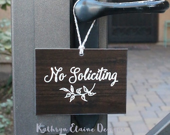 以下が含まれることがあります： 白い文字で「No Soliciting」と書いてある、濃い茶色の木製の看板。白い花の模様が描かれている。看板は白い紐で黒いドアノブにかけられている。