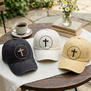 May include: Three baseball caps in black, off-white, and tan, each featuring a circular emblem with a cross in the center. The black cap has a light-colored cross, while the off-white and tan caps display a dark cross within the circle.