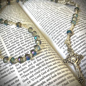 May include: A rosary with iridescent blue and silver beads rests on an open book. The silver crucifix features the figure of Jesus. The book's pages display printed text.