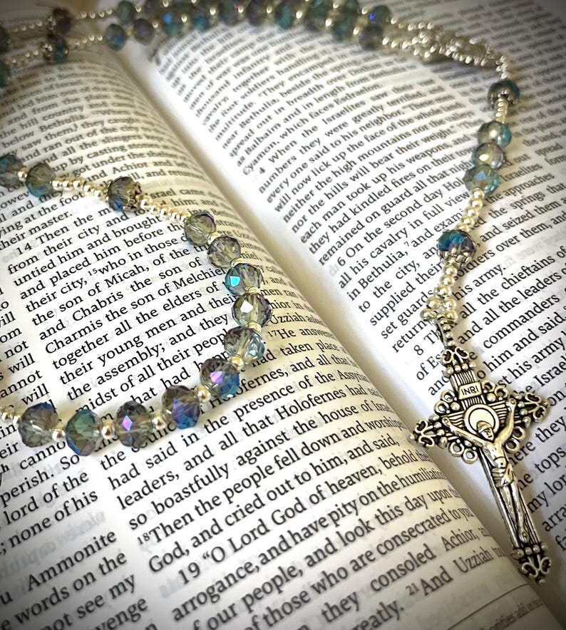 May include: A rosary with iridescent blue and silver beads rests on an open book. The silver crucifix features the figure of Jesus. The book's pages display printed text.