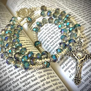 May include: A rosary with iridescent blue and green beads, resting on an open book. The rosary features a silver crucifix with the inscription "INRI". The beads are faceted and reflect light.