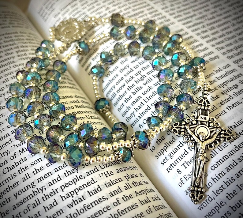 May include: A rosary with iridescent blue and green beads, resting on an open book. The rosary features a silver crucifix with the inscription "INRI". The beads are faceted and reflect light.