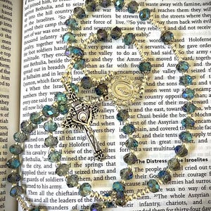 May include: A rosary with iridescent blue and green beads, a silver crucifix, and a detailed centerpiece. The rosary is displayed on an open book with religious text.