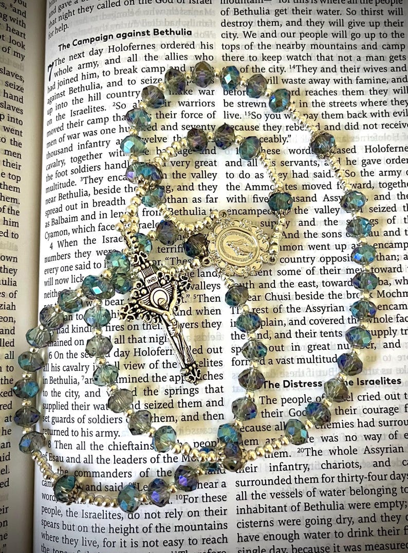 May include: A rosary with iridescent blue and green beads, a silver crucifix, and a detailed centerpiece. The rosary is displayed on an open book with religious text.
