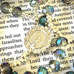 May include: A rosary with iridescent blue and gray faceted beads and a silver-toned centerpiece. The centerpiece features a religious emblem. The rosary is set against a backdrop of printed text.