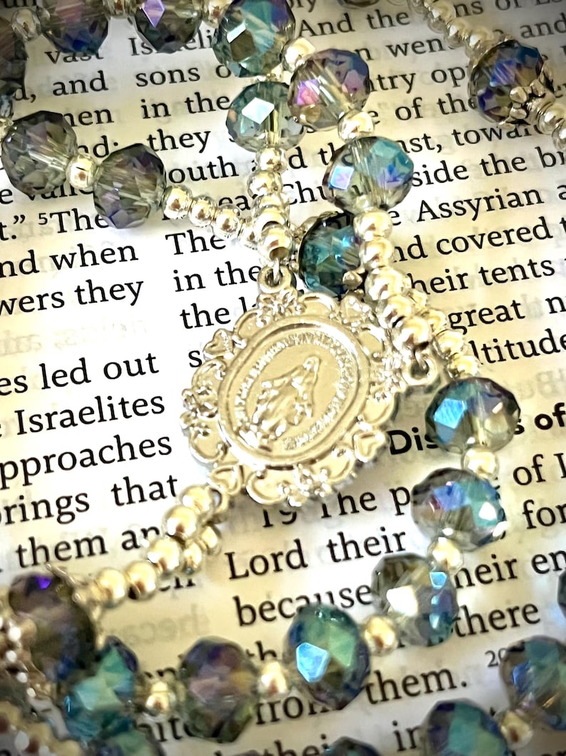 May include: A rosary with iridescent blue and gray faceted beads and a silver-toned centerpiece. The centerpiece features a religious emblem. The rosary is set against a backdrop of printed text.