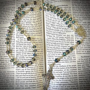May include: A rosary with iridescent beads and a silver-toned crucifix lies open on a book. The beads are a mix of blues and greens. The book's pages are yellowed, with text visible.