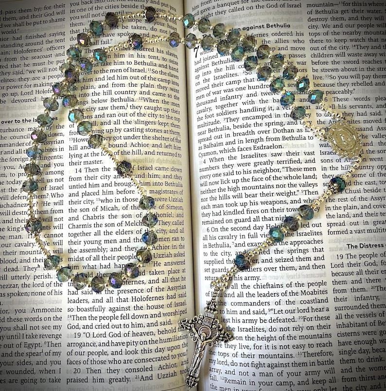 May include: A rosary with iridescent beads and a silver-toned crucifix lies open on a book. The beads are a mix of blues and greens. The book's pages are yellowed, with text visible.