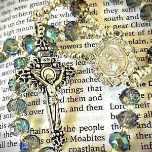 May include: A rosary with iridescent blue and silver beads. The silver crucifix features the figure of Jesus, and the centerpiece has a religious emblem. The rosary is set against a backdrop of open book pages.