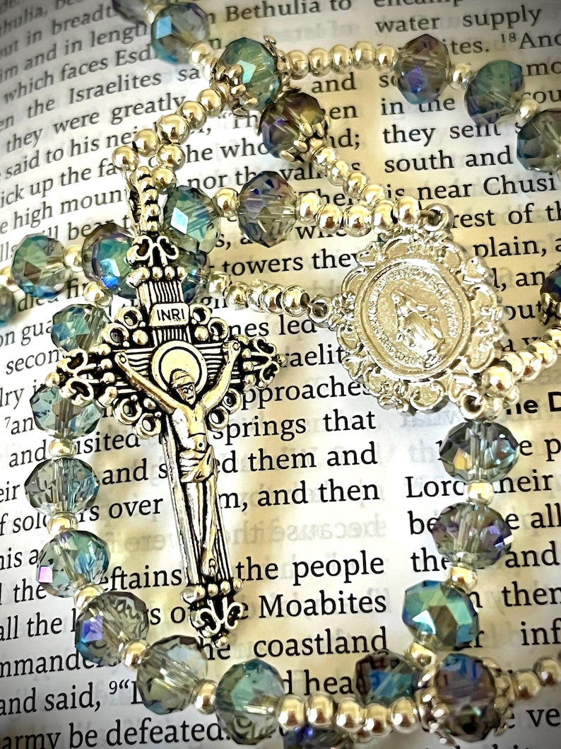 May include: A rosary with iridescent blue and silver beads. The silver crucifix features the figure of Jesus, and the centerpiece has a religious emblem. The rosary is set against a backdrop of open book pages.